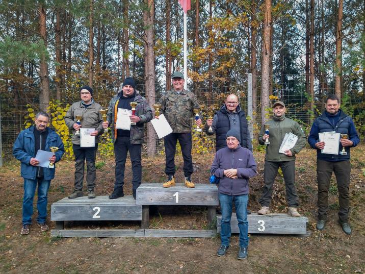 Zdjęcie&#x20;przedstawia&#x20;grupę&#x20;mężczyzn&#x20;stojących&#x20;na&#x20;podium&#x20;w&#x20;lesie&#x2c;&#x20;trzymających&#x20;puchary&#x20;i&#x20;dyplomy&#x2e;&#x20;To&#x20;zwycięzcy&#x20;XI&#x20;Zawodów&#x20;Strzeleckich&#x20;o&#x20;Puchar&#x20;Nadleśniczego&#x20;Nadleśnictwa&#x20;Giżycko&#x2e;&#x20;Na&#x20;pierwszym&#x20;miejscu&#x20;stoi&#x20;mężczyzna&#x20;w&#x20;ubraniu&#x20;moro&#x2c;&#x20;na&#x20;drugim&#x20;i&#x20;trzecim&#x20;–&#x20;inni&#x20;uczestnicy&#x20;z&#x20;pucharami&#x2e;&#x20;Pozostali&#x20;stoją&#x20;po&#x20;bokach&#x20;podium&#x2e;&#x20;W&#x20;tle&#x20;widoczne&#x20;są&#x20;drzewa&#x20;i&#x20;ogrodzenie&#x2e;